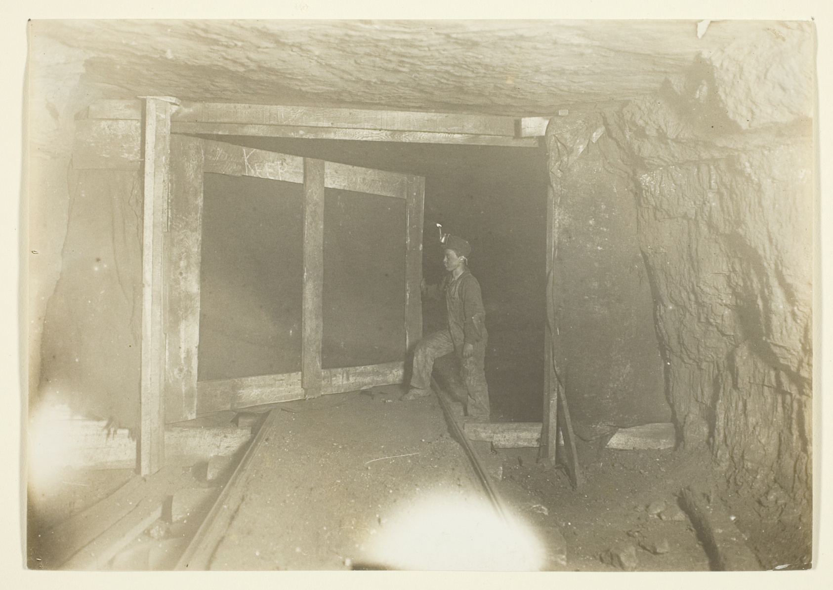 A timbered mine tunnel photographed head-on, rails running away into the dark, with the small figure of a boy standing beside the ventilation door at the far end.