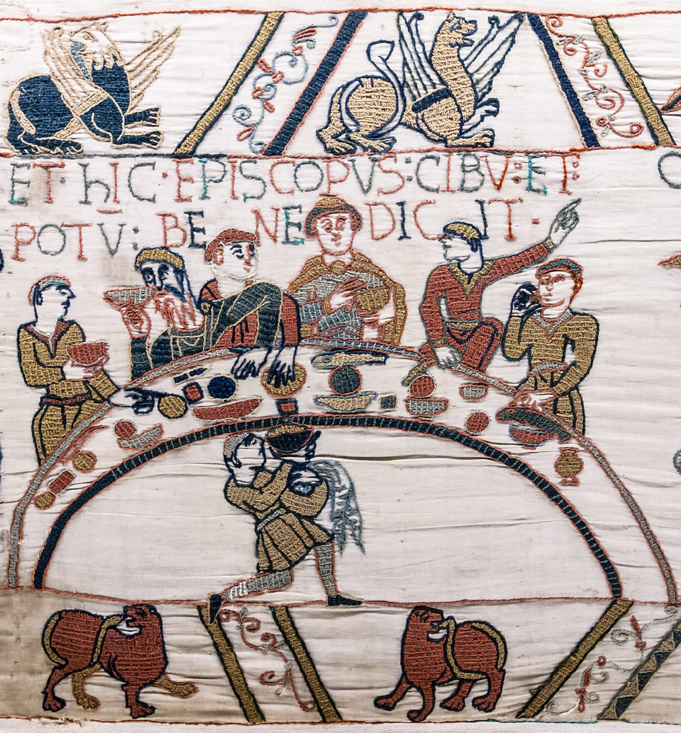 Embroidered banquet scene from the Bayeux Tapestry: figures seated behind a curved table laden with food and drink, with Bishop Odo raising his hand in blessing at the centre.