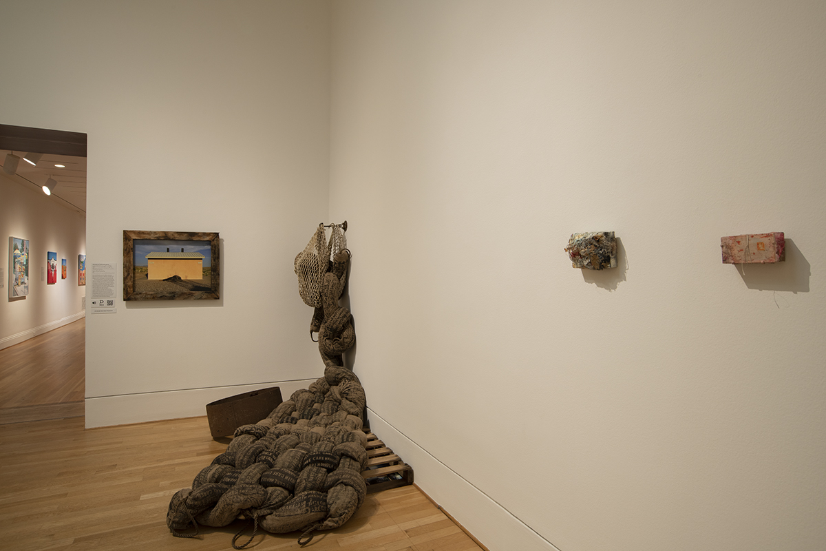 Gallery corner with a large woven floor sculpture and small wall-mounted works