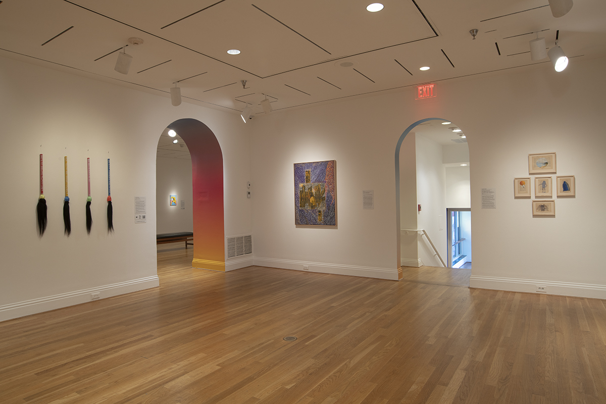 Gallery with hanging broom-like sculptures of human hair and plastic rattan, framed works on paper, and two arched doorways