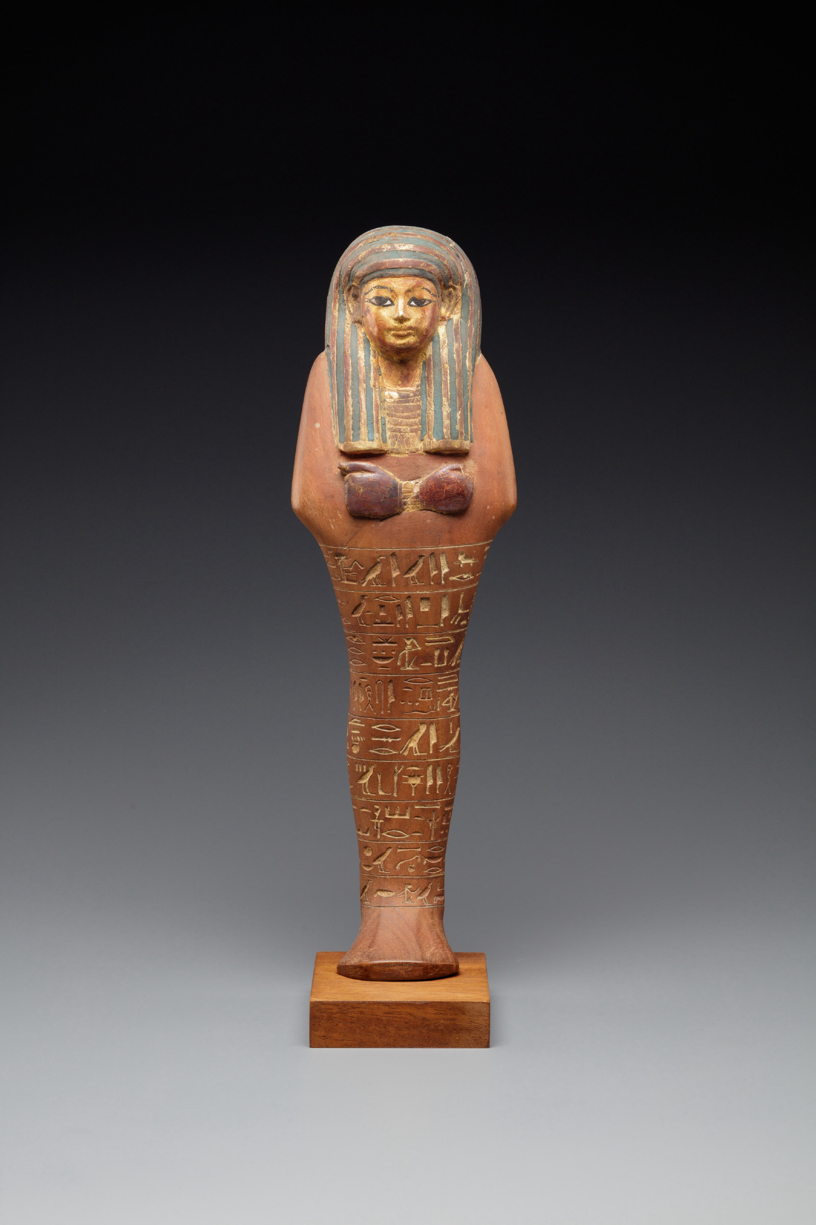 A small mummiform shabti figure standing on a base, with a painted face, a blue-striped wig falling to the shoulders and columns of hieroglyphs down the front of the body.