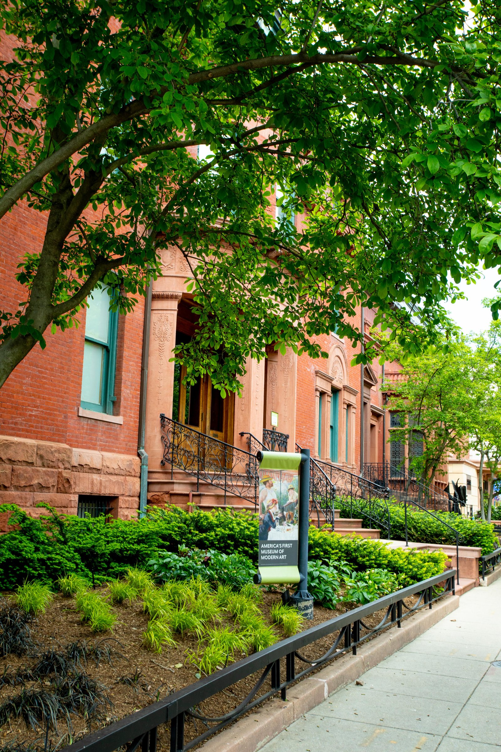 Red-brick Phillips Collection building seen from the sidewalk through leafy trees, with an exhibition banner