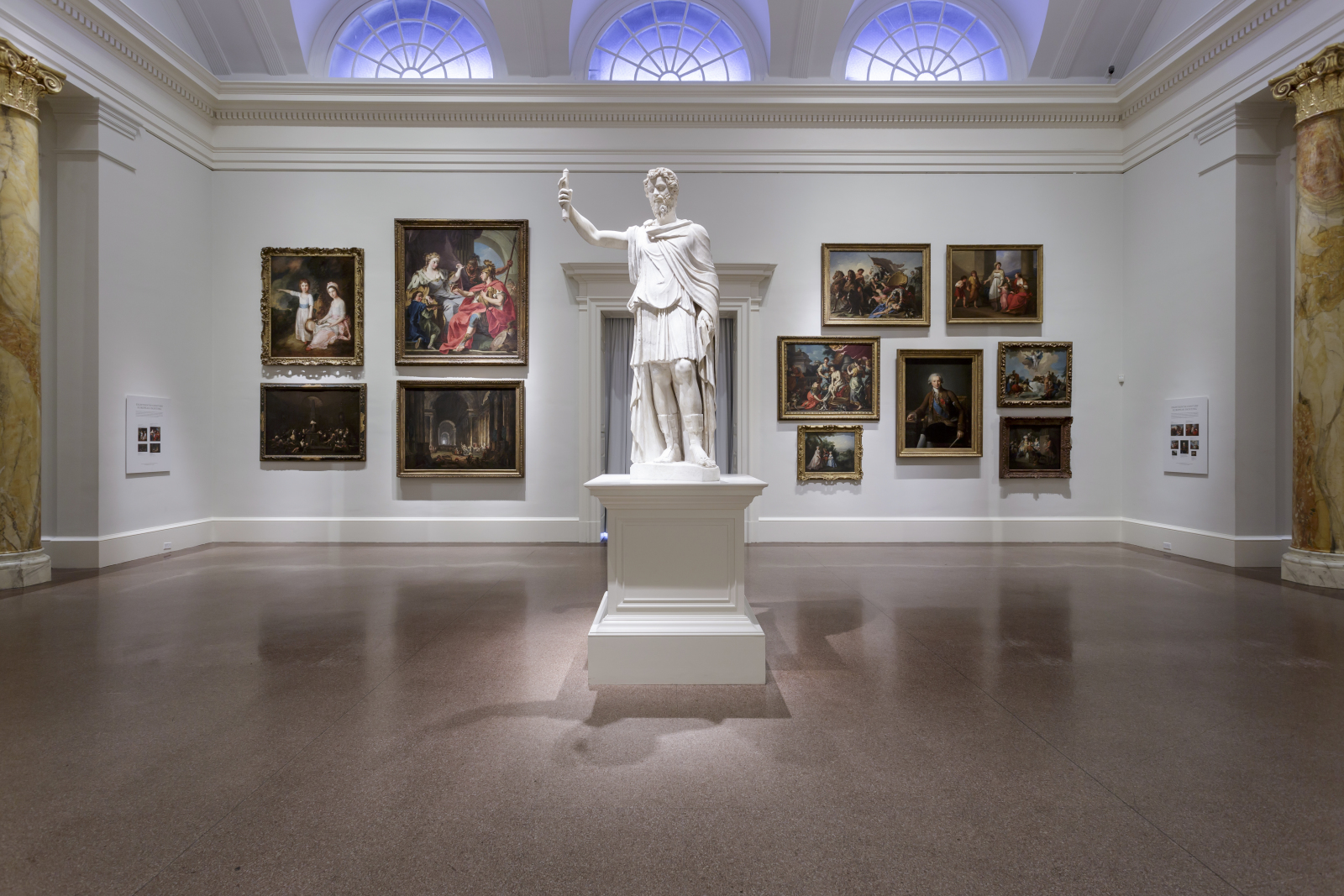 Neoclassical gallery with a white marble statue on a plinth at its centre, old master paintings hung in two tiers on pale walls, and arched lunette windows above
