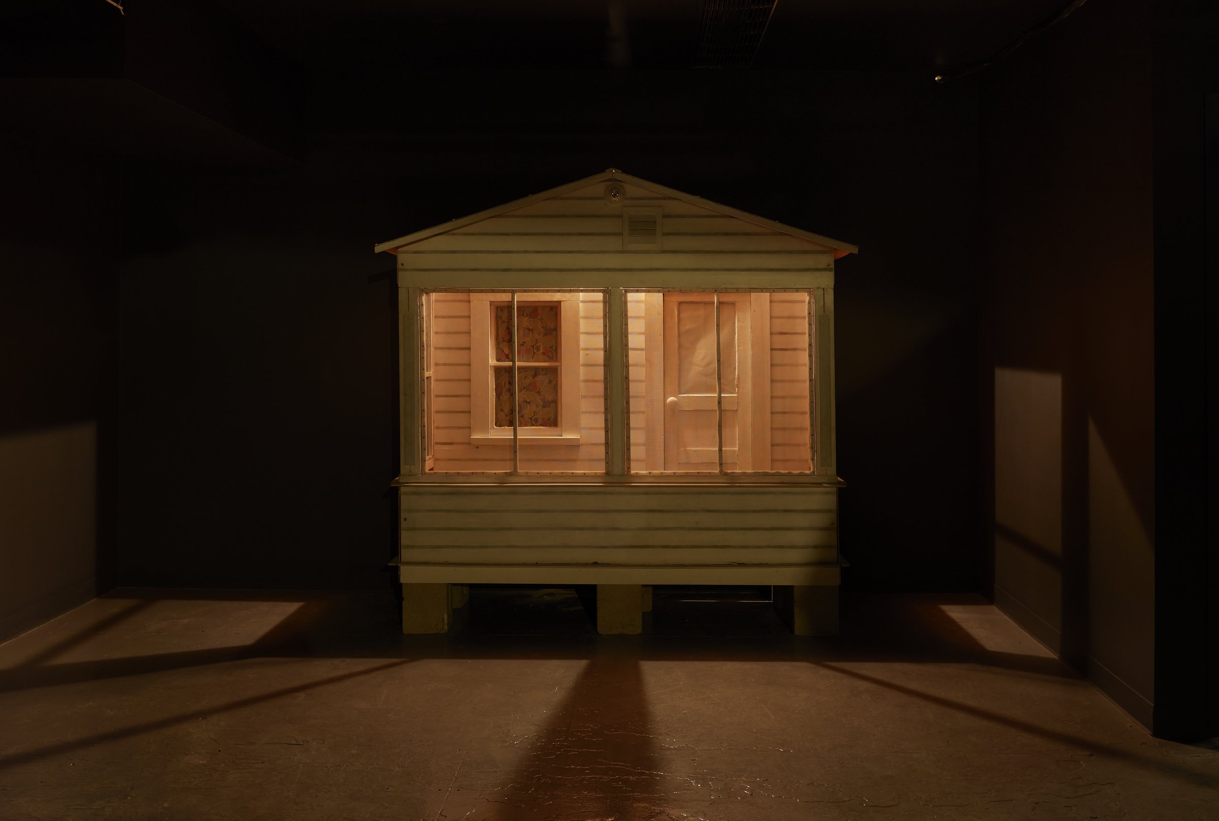 Donna Dennis sculpture of a small lit tourist cabin porch glowing in a dark gallery