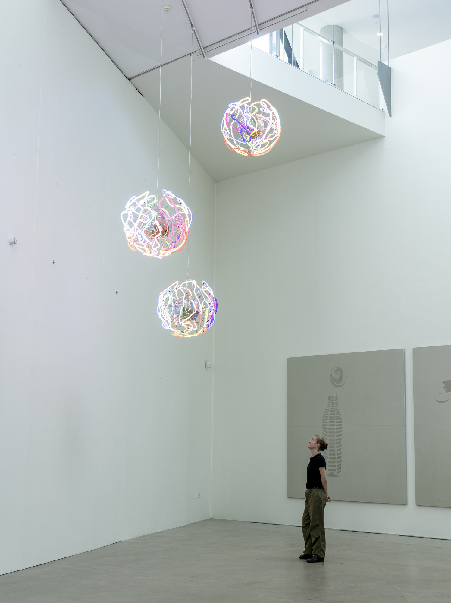 Three illuminated spherical sculptures made of tangled colored neon-like lines, suspended from the ceiling with a visitor looking up