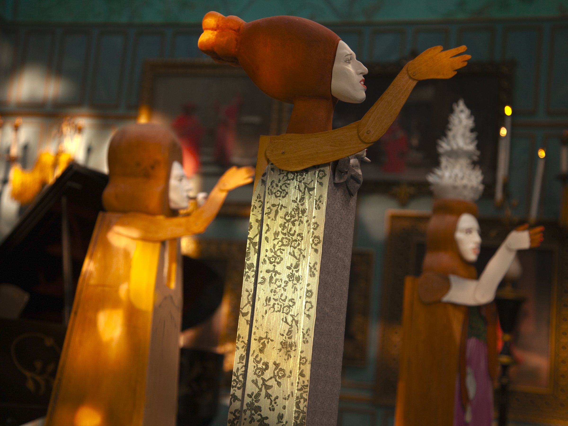 Stop-motion still: tall puppet figures with pale masks raise their arms in a dim, candlelit room