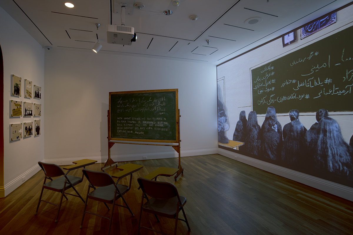 Dimly lit gallery with school desk chairs facing a projected video of a chalkboard and a row of seated figures
