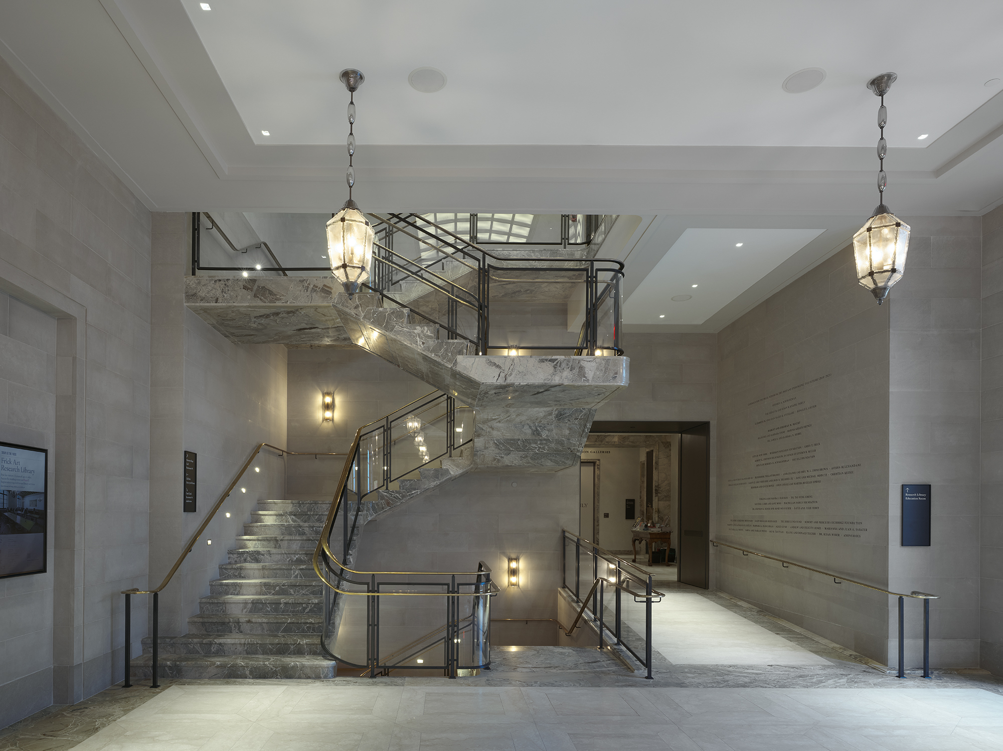 The James S. and Barbara N. Reibel Reception Hall, part of the renovated Frick: a pale stone hall with a sculptural staircase and pendant lamps.