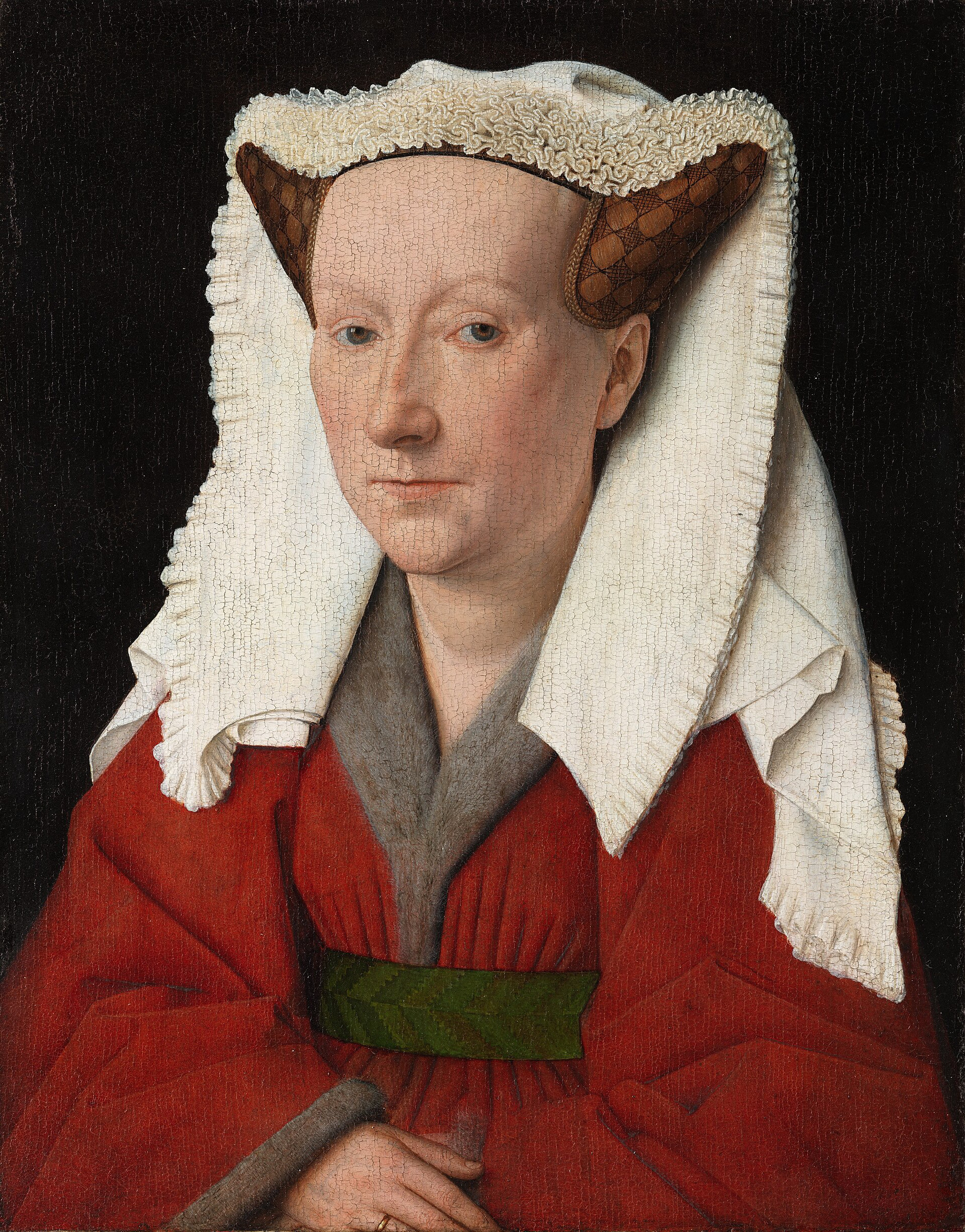 Portrait of a woman in a red dress trimmed with grey fur, her hair drawn up into a horned headdress of pleated white linen, set against a dark background with inscriptions on the painted frame.