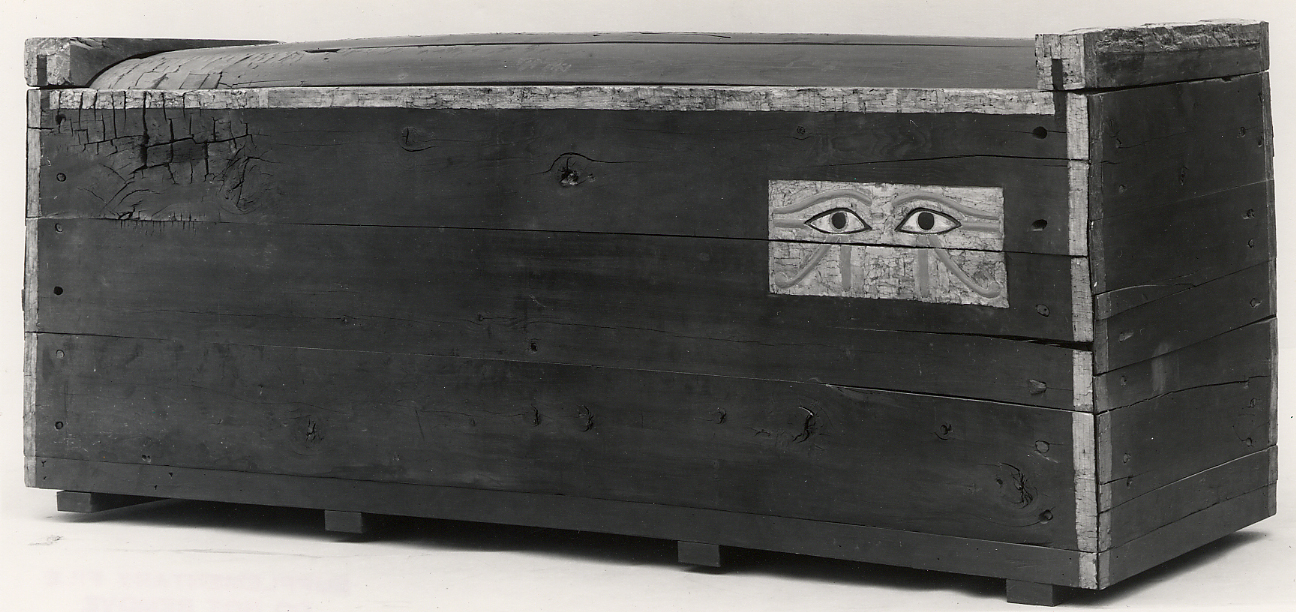 A plain rectangular wooden coffin from Middle Kingdom Egypt, the dark boards bare except for a small painted panel of two eyes near one end.