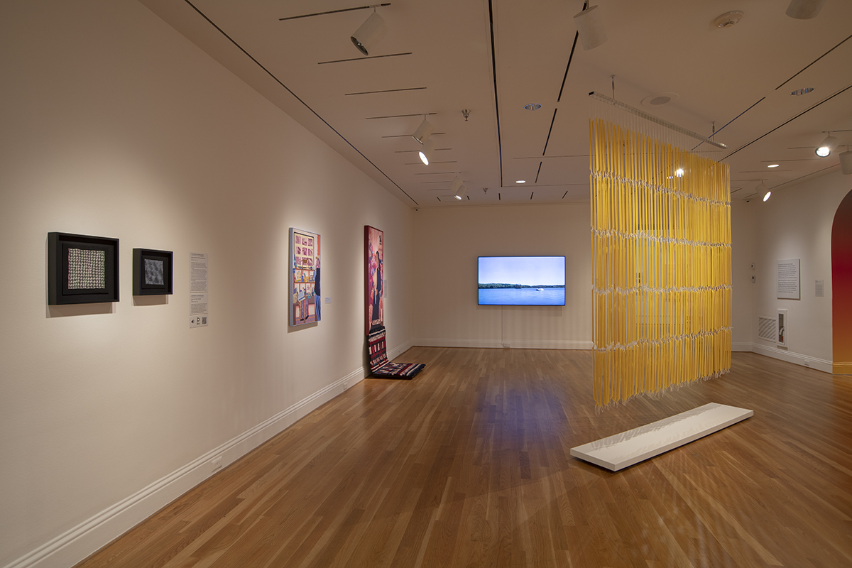Gallery hung with a curtain of gold vertical strands, framed paintings along the wall and a video monitor beyond