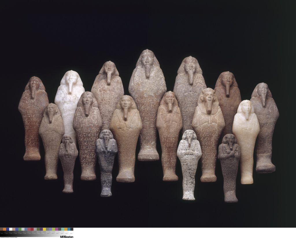 Frontal view of 17 stylized figures of various sizes with headdresses, long, narrow beards, and writing on their torsos and legs.