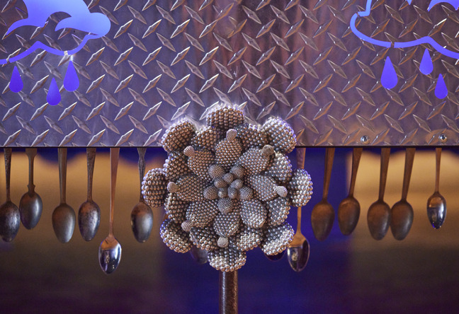 Detail of a large metal flower sculpture before a row of hanging spoons against a blue-lit wall