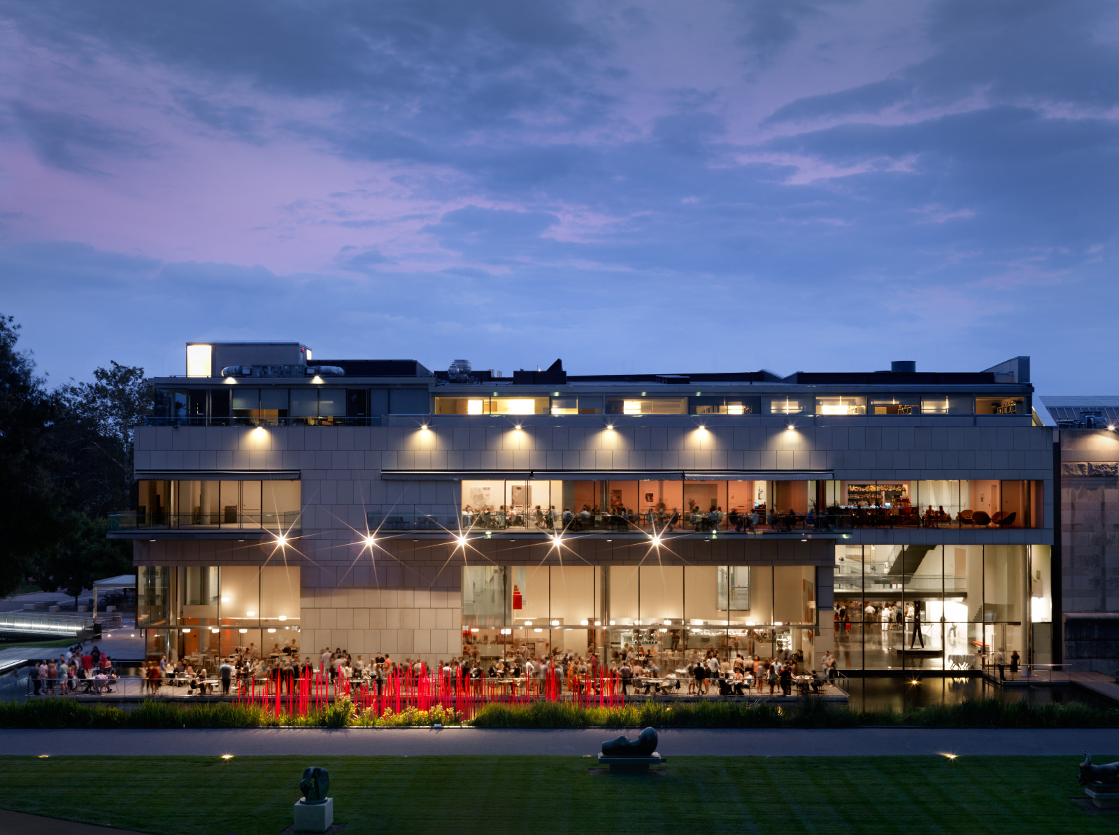 The James W. and Frances G. McGlothlin Wing lit from within at dusk, its glass and stone façade above a terrace of diners, with lawn in the foreground and a violet twilight sky