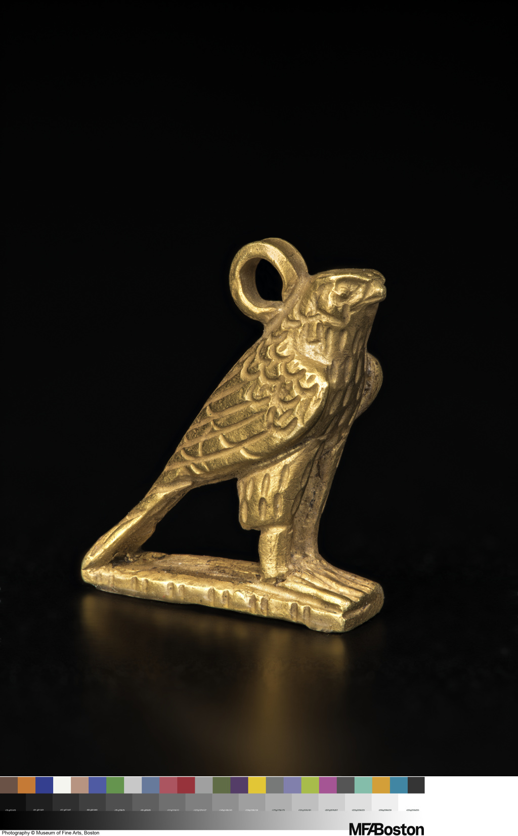 Amulet of a standing gold bird with folded wings, tips touching the ground. A ring is attached to its head.