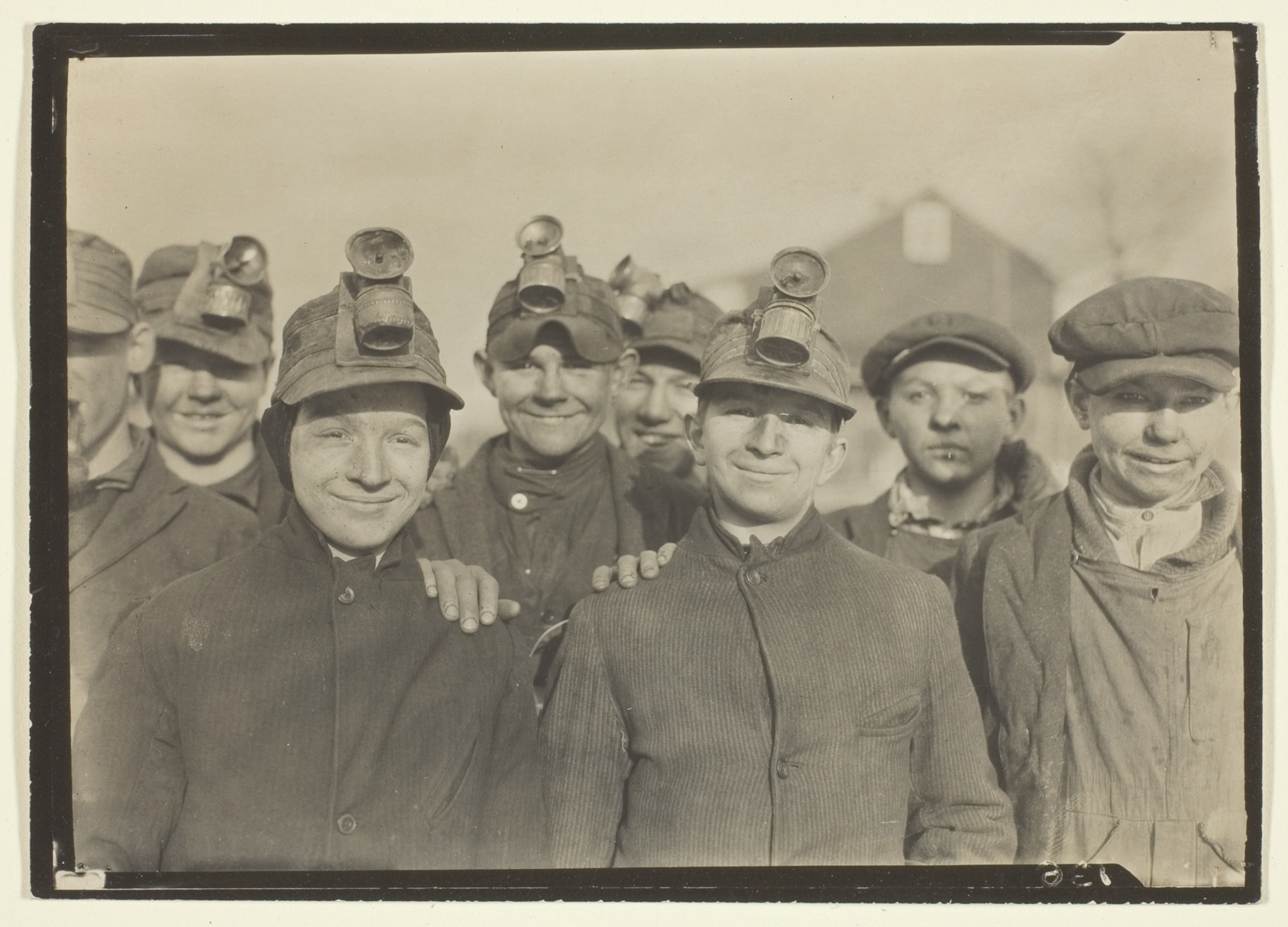A close group portrait of eight miners and boys pressed shoulder to shoulder, wearing peaked caps fitted with lamps, their faces and clothes dark with coal dust as they look at the camera.