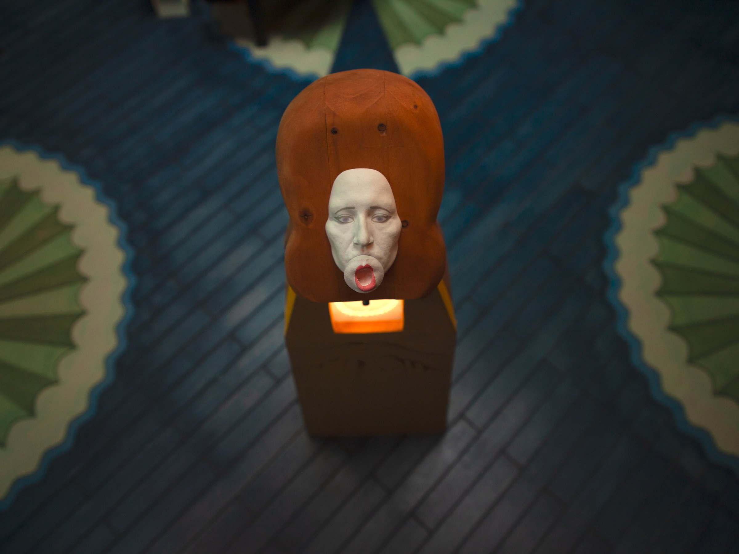 Stop-motion still: a single masked puppet with red hair holding a candle on a dark patterned floor