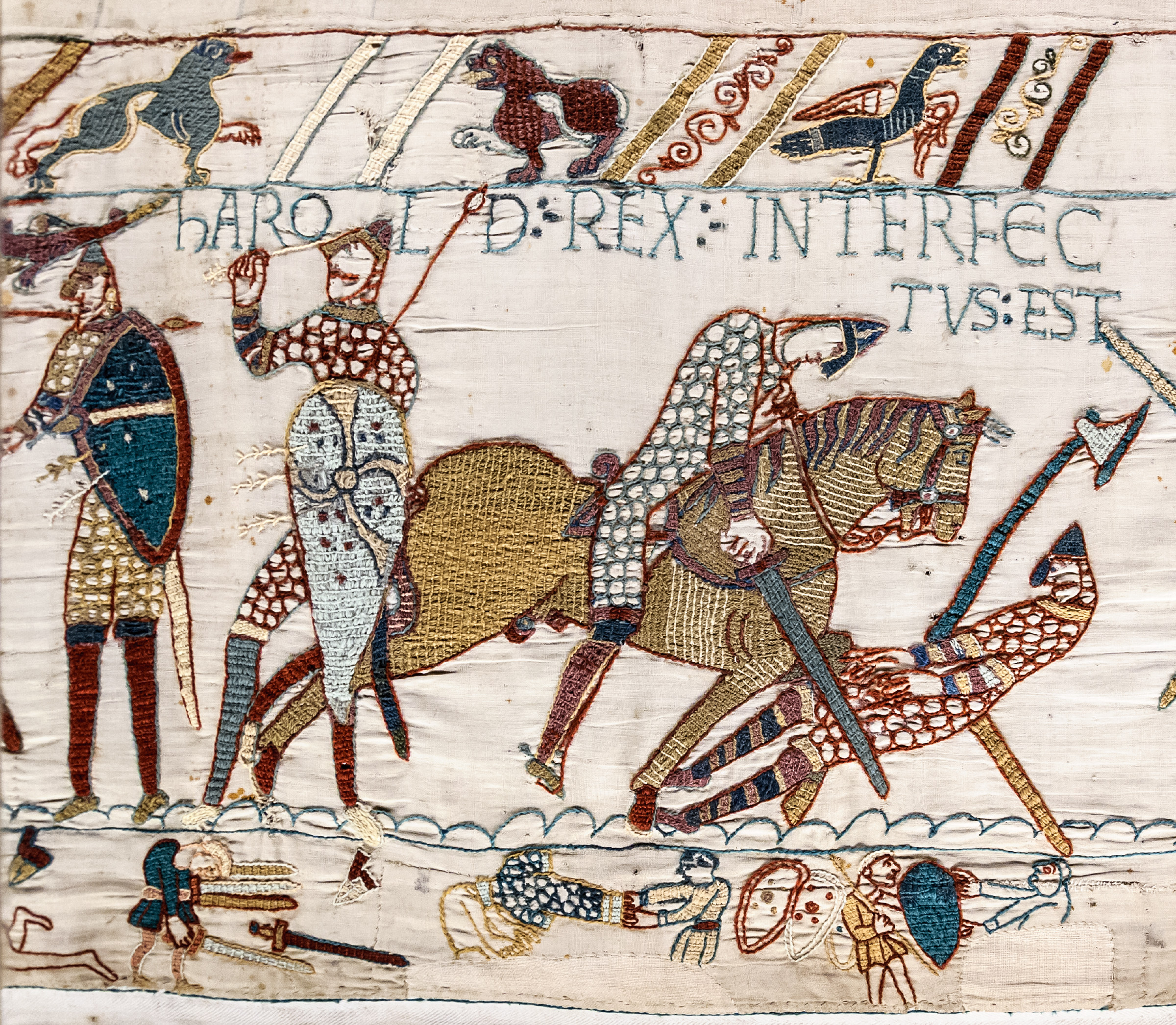 Embroidered scene from the Bayeux Tapestry showing mounted Norman knights riding down English foot soldiers, with the Latin inscription naming Harold's death worked into the upper border.