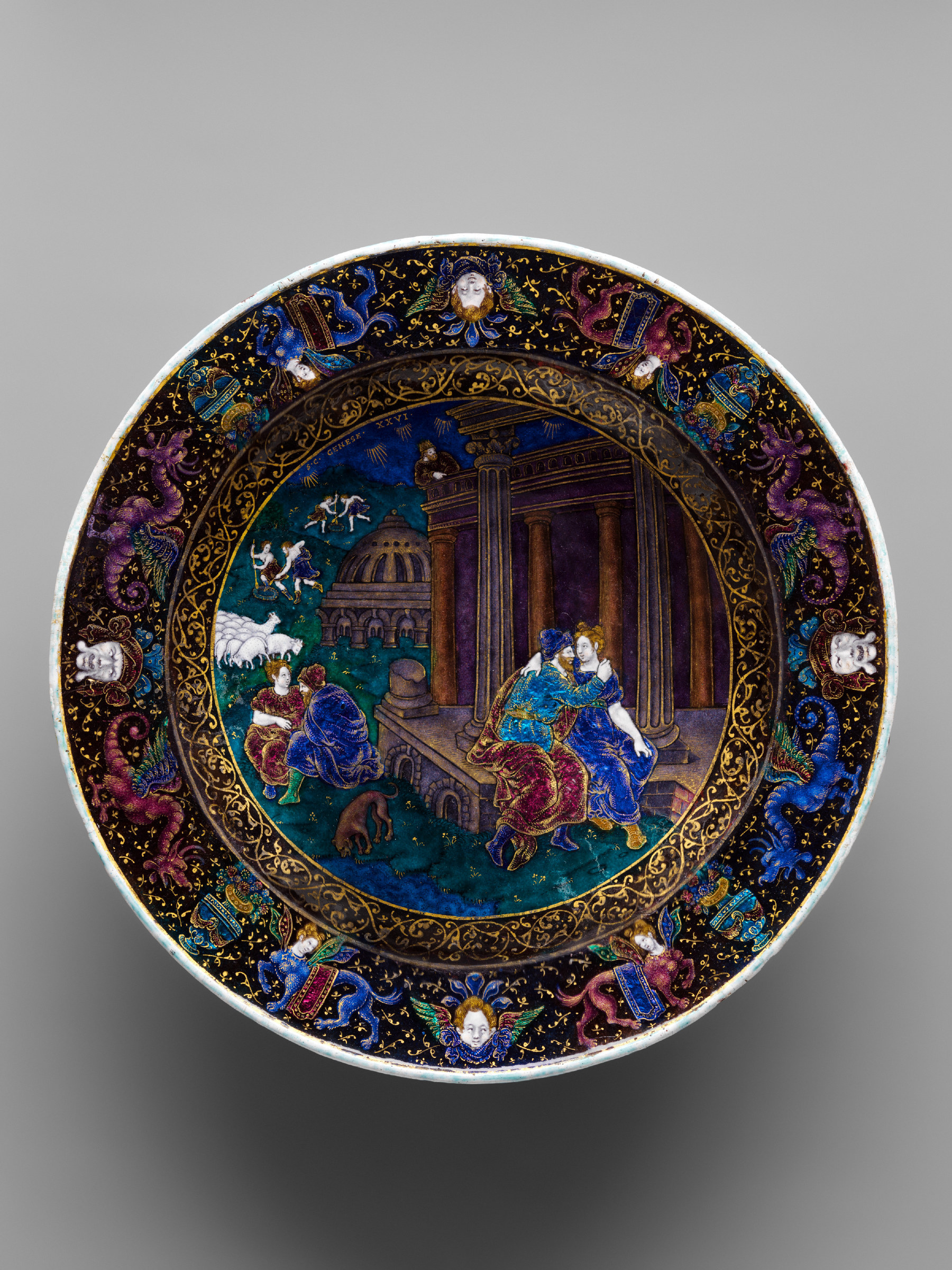 A circular painted enamel dish on copper: a scene of figures in an interior at the centre, surrounded by a deep border of scrolling ornament in blue, red and gold set with pale jewel-like bosses.