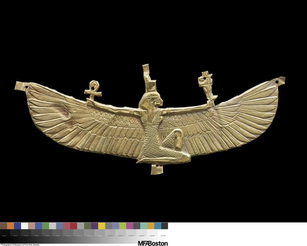 Flat gold pectoral of a kneeling woman wearing a tall headdress. She stretches out winged arms and holds two implements.