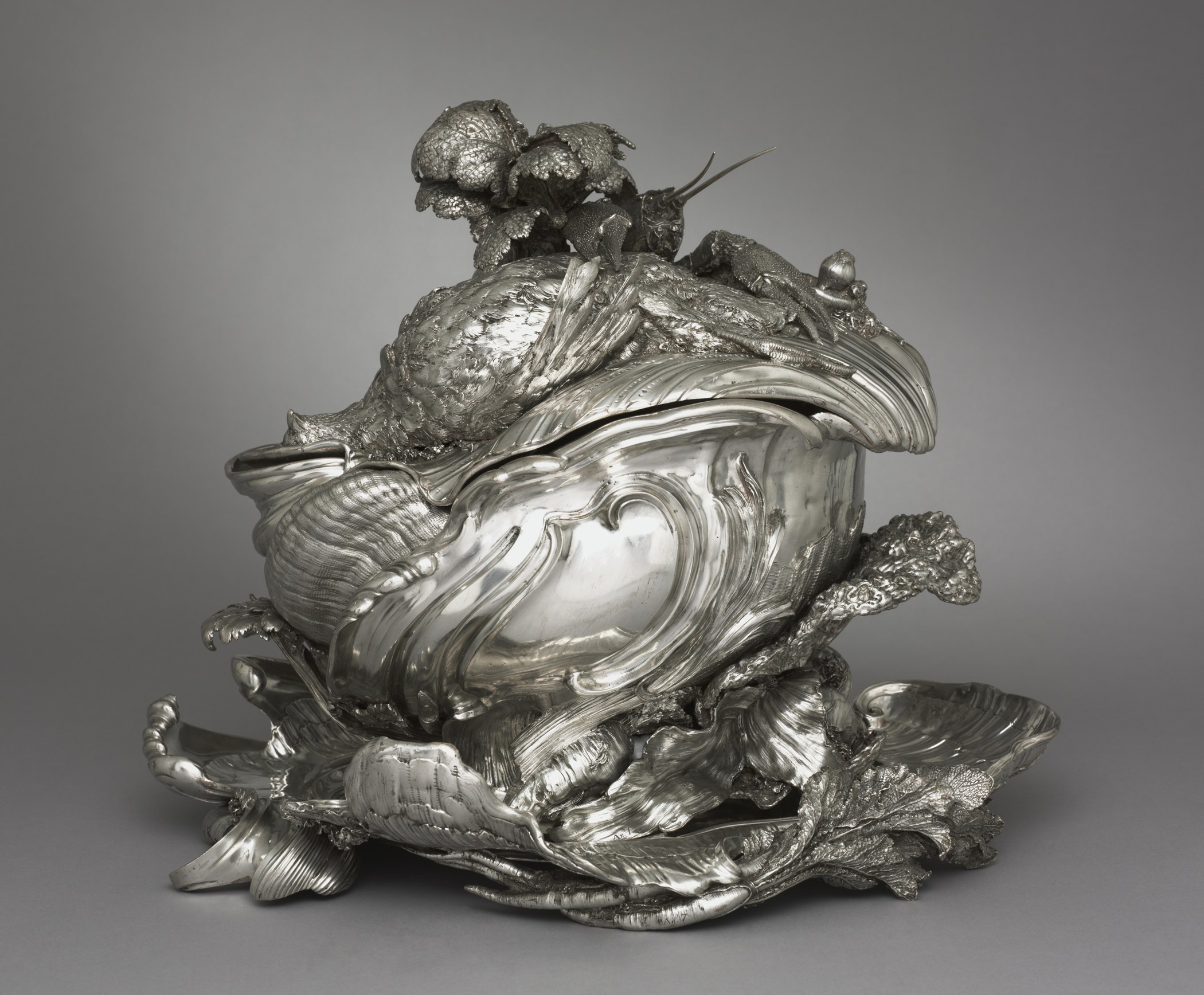 Rococo silver tureen on a stand, its swirling shell-like body covered with cast vegetables, a fish and game
