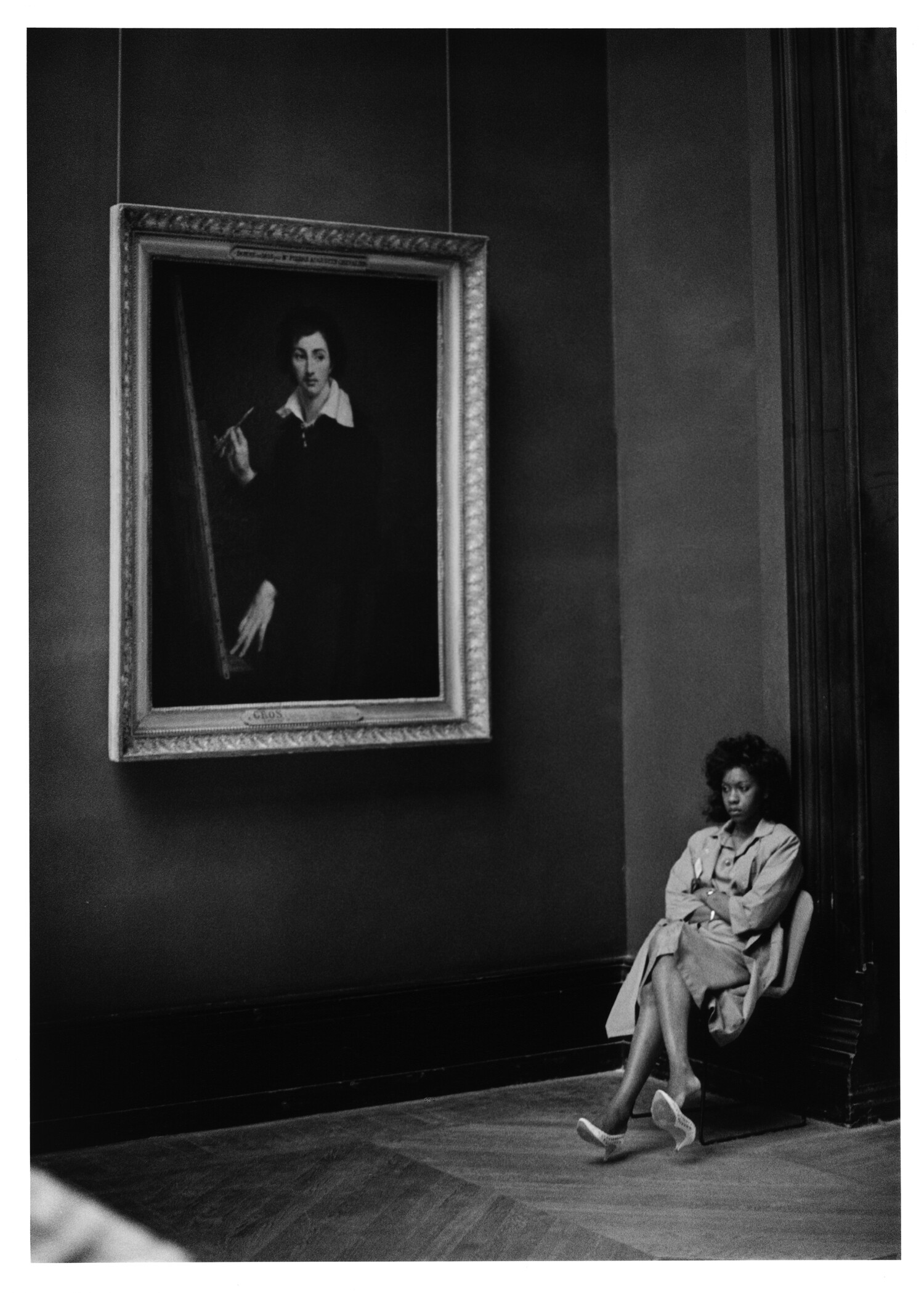 Black and white photograph of a woman seated alone on a bench in a Louvre gallery beneath a large framed portrait of a painter holding a brush
