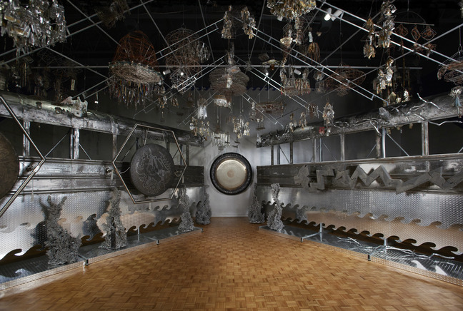Steel frame installation hung with gongs, horse figures and wire objects above a wooden floor