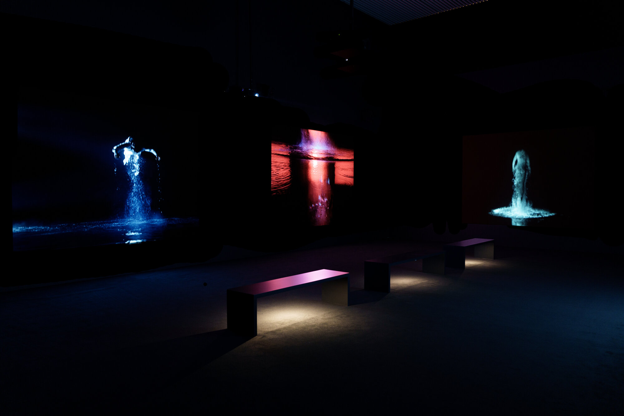 Dark video installation gallery with three screens showing glowing blue, red, and teal figures in water, with benches for viewing