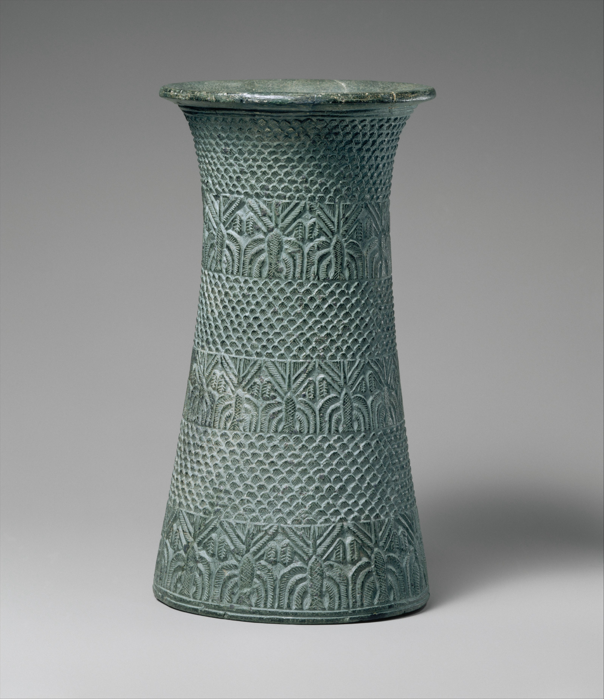 A tall cylindrical stone vessel with a flared rim, carved all over in low relief with an overlapping scale pattern, the surface a mottled grey-green.