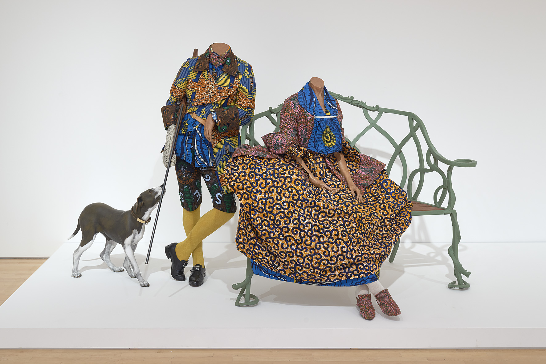 Sculpture of two headless mannequins dressed in brightly patterned Dutch wax textiles, posed as an eighteenth-century couple beside a green iron bench with a model dog