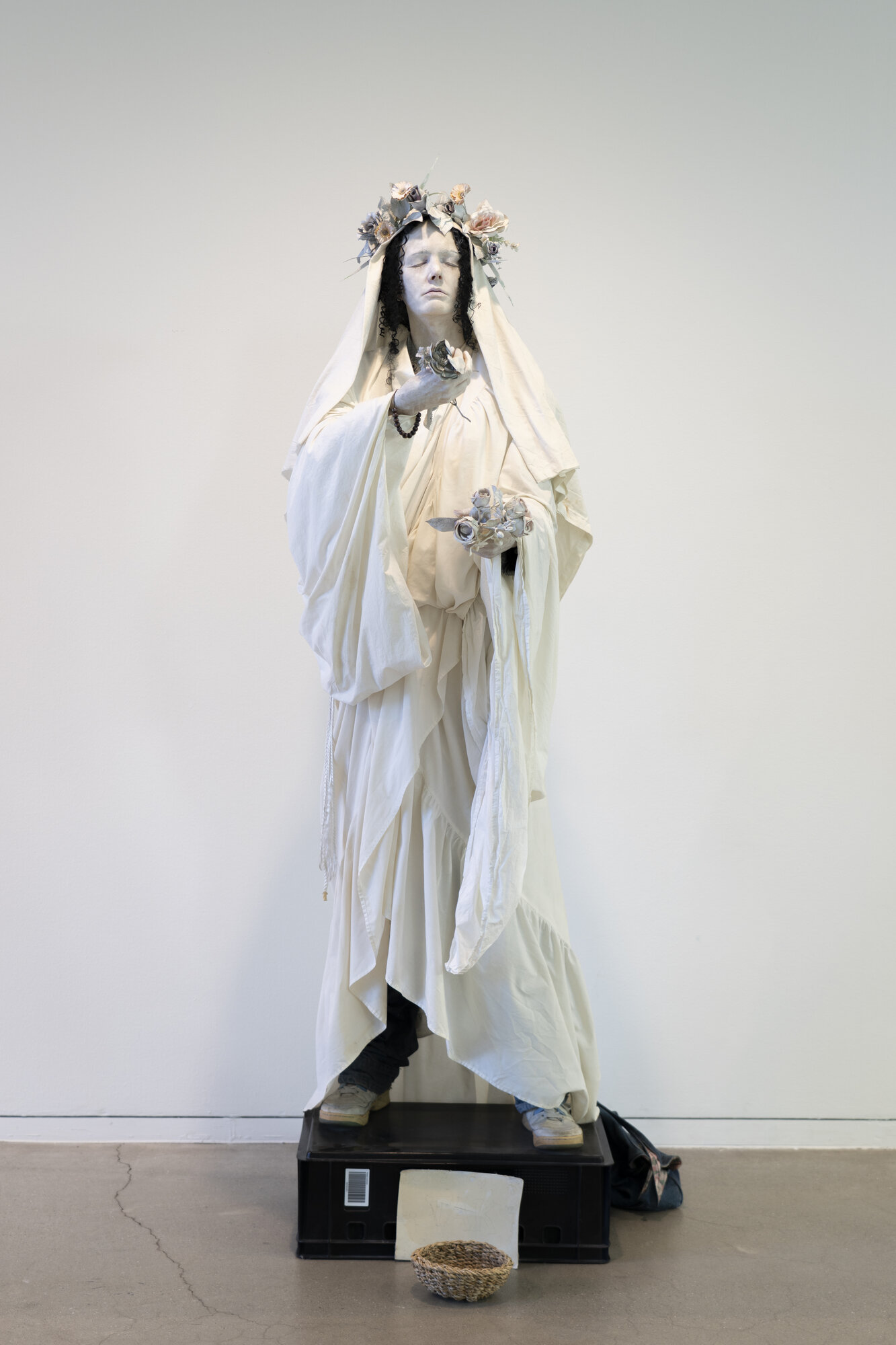Ghostlike sculpture of a veiled figure in draped white fabric with a floral wreath, standing on a black crate with an offering bowl