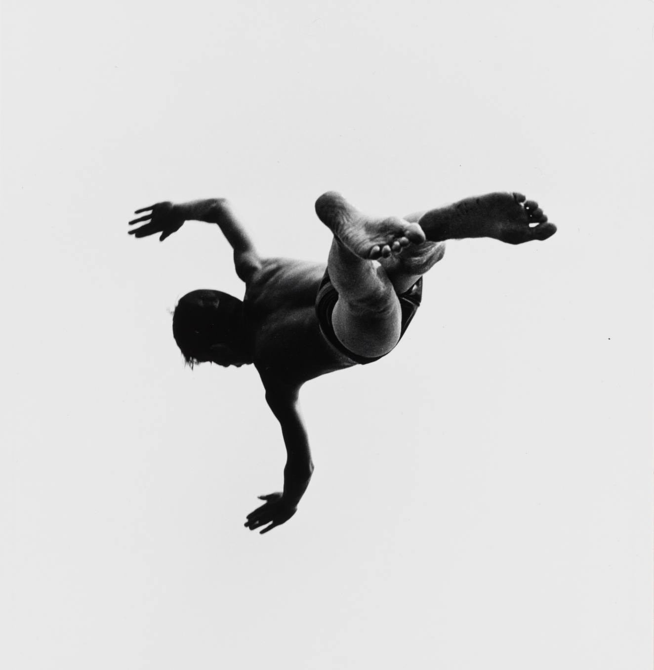 Black and white photograph of a male figure that could be flying or falling through the air, arms outspread and face in profile, dramatically contrast against a white sky.