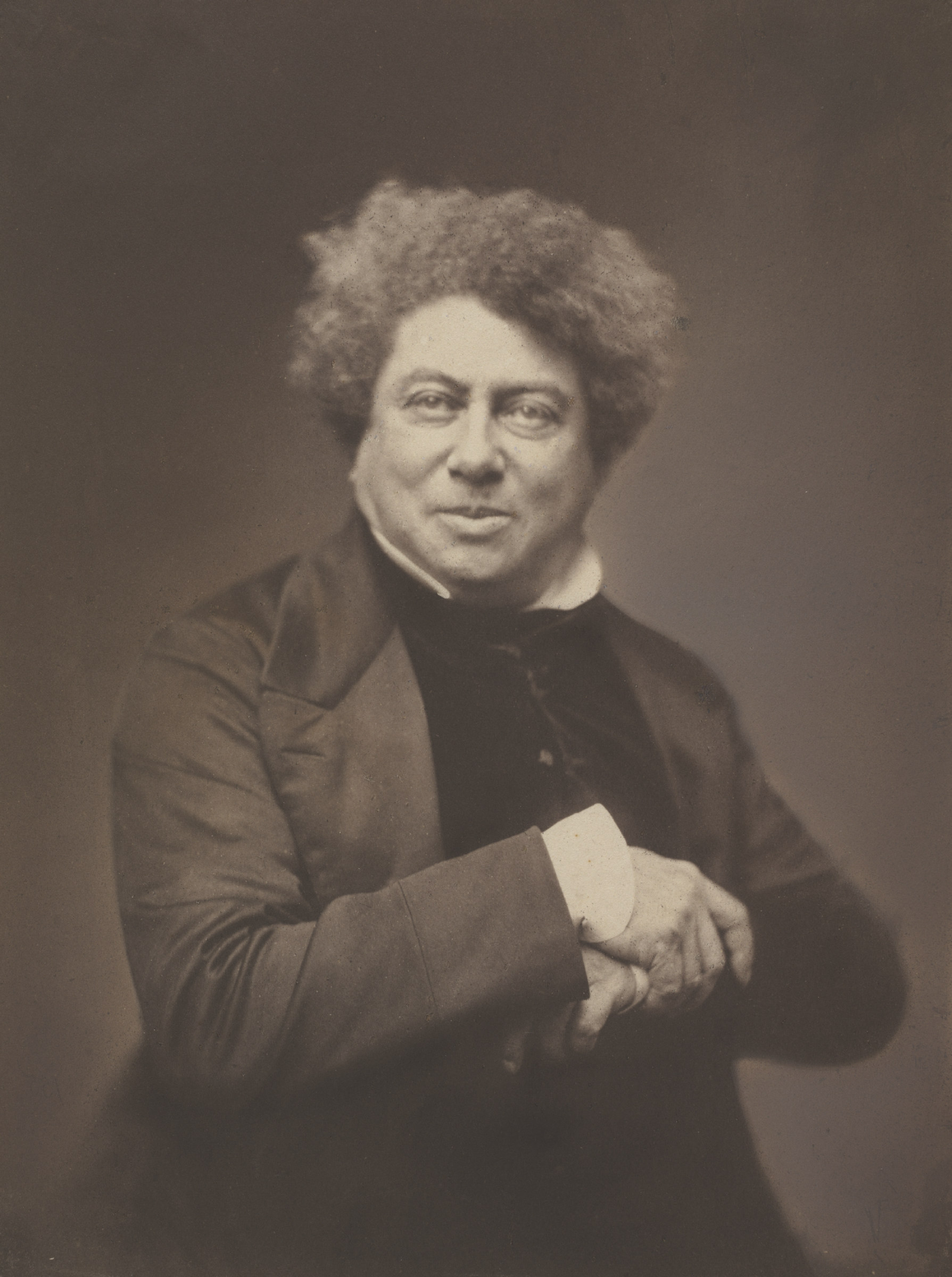 A salted paper portrait of Alexandre Dumas seated with his hands clasped in his lap, his thick curling hair and heavy features lit from one side against a plain backdrop.