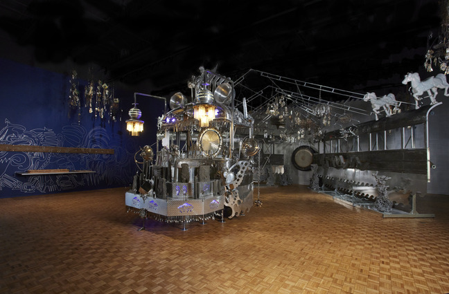 Wide installation view with Maravilla altar at left and a long metal wall of hanging objects and horse figures