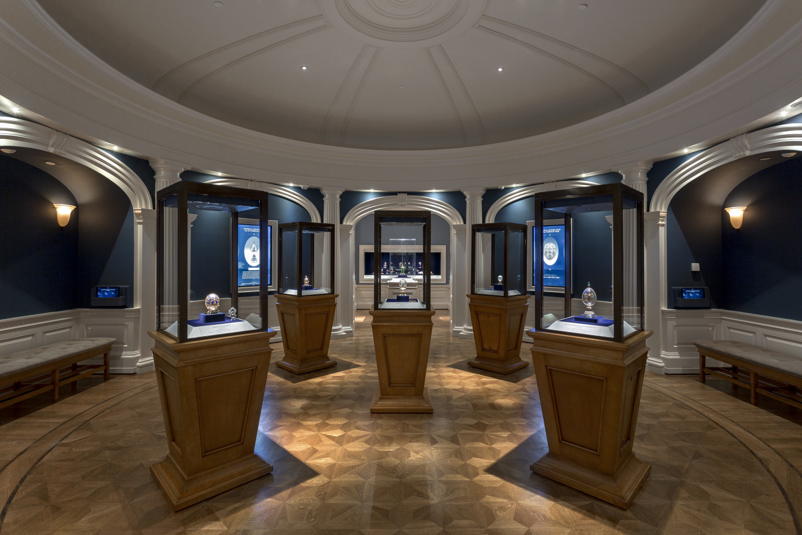Domed circular gallery with deep blue alcoves, illuminated glass display cases on wooden plinths ringing the room, and benches between them