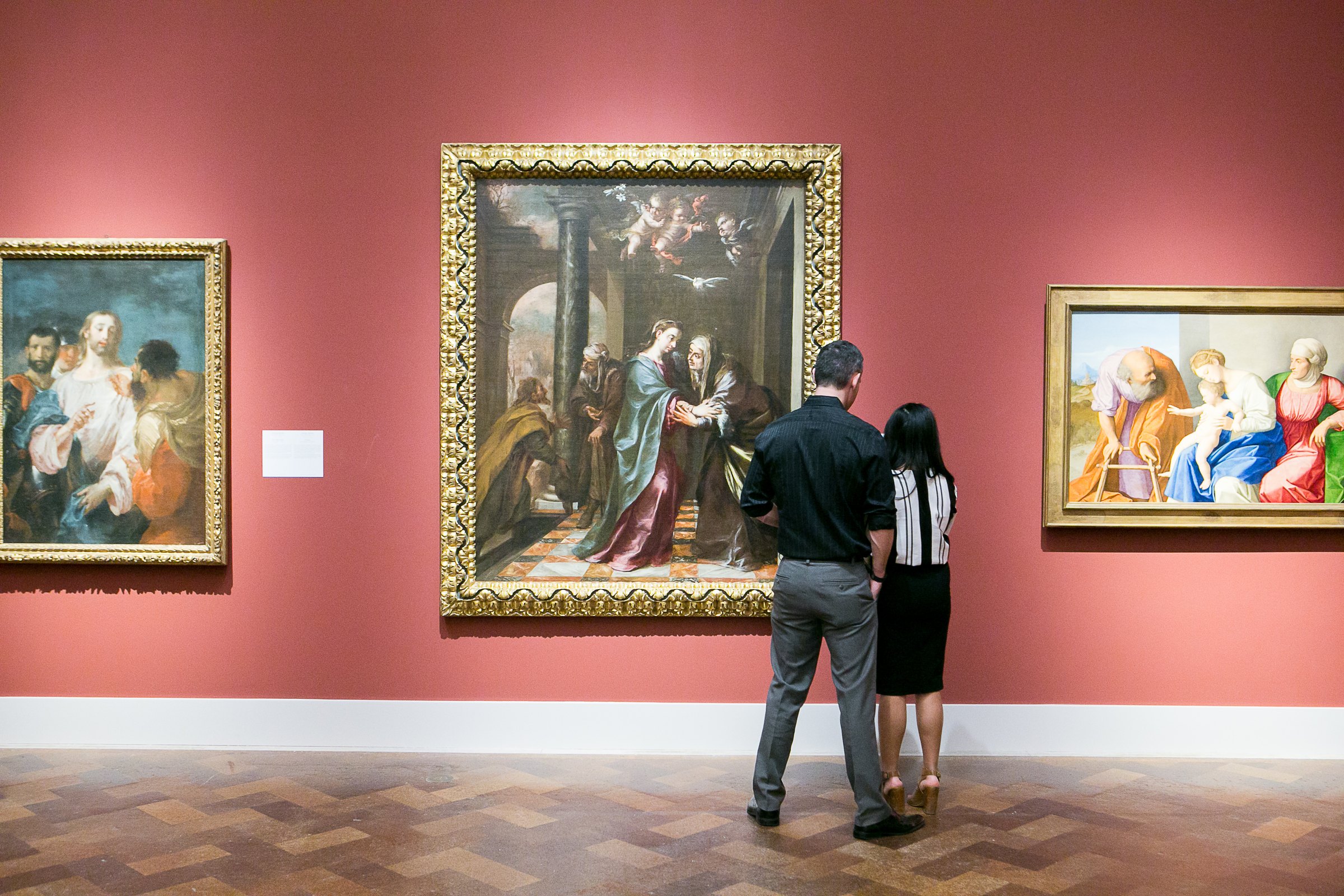Two visitors looking at a large altarpiece in a red-walled gallery